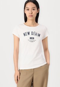 T-shirt bianca a maniche corte con la scritta "NEW DENIM CLUB" stampata in blu navy, scollo rotondo e una texture morbida, abbinata a pantaloni beige.