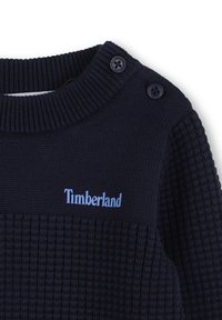 Granatowy dziany sweter z teksturowanym wzorem, z prążkowanym kołnierzem, dwoma niebieskimi guzikami na ramieniu oraz logo "Timberland" w jasnoniebieskim kolorze.