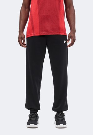 Mann, der schwarze Under Armour Jogginghosen, schwarz-weiße Sneakers und ein rotes Sportshirt trägt, vor weißem Hintergrund stehend.