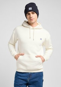 Light cream hoodie with a front pocket, drawstrings, and a small logo. Worn with a dark blue ribbed beanie and blue denim jeans.