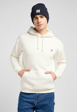 Light cream hoodie with a front pocket, drawstrings, and a small logo. Worn with a dark blue ribbed beanie and blue denim jeans.