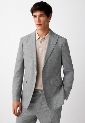 Grau taillierter Blazer mit Einsätzen an den Revers, zwei Fronttaschen und passender Hose. Getragen über einem hellbeigen Strick-Polohemd mit Knöpfen.