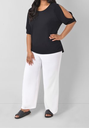 Kobieta ubrana w czarną bluzkę z odkrytymi ramionami, białe szerokie spodnie, czarne klapki, stojąca z jedną ręką na biodrze na jednolitym tle.