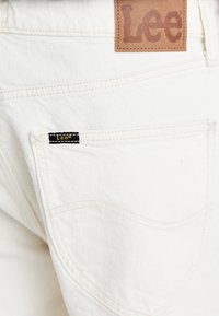 Jeans en denim blanc avec une étiquette en cuir Lee au niveau de la taille et une petite étiquette noire sur la poche arrière. Présente des coutures et un design de poche classique.