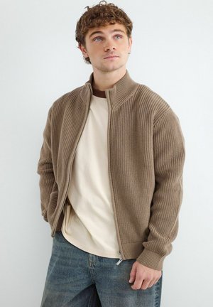 Junger Mann mit lockigem Haar trägt einen beigen gestrickten Reißverschluss-Pullover über einem weißen Hemd und blauen Jeans, schaut leicht nach oben.