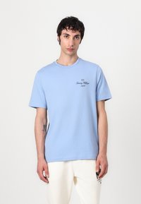 T-shirt en coton bleu clair à manches courtes, col rond et petit logo noir imprimé sur la poitrine. Texture douce et coupe décontractée.