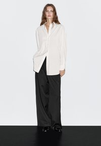 Camicia bianca con bottoni, a righe verticali, abbinata a pantaloni neri larghi. Tessuto morbido e vestibilità comoda, con colletto e polsini classici.