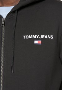 Sweat à capuche zippé noir avec le texte blanc "TOMMY JEANS" et un petit logo rouge, blanc et bleu. Tissu texturé avec capuche à cordon.