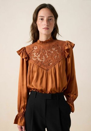 Rust-colored satin blouse with lace detailing on the bodice, ruffled sleeves, and a high neck. Paired with black tailored trousers.