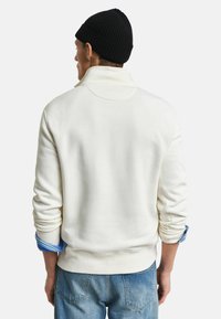 Crème-kleurige sweatshirts met een hoge kraag, ribgebreide manchetten en een gladde textuur. Gemaakt over een blauwe overhemd, gecombineerd met lichtblauwe jeans.