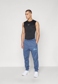 Nike sort tanktop, blå svedpants med logo og hvide sneakers. Materialet ser glat ud; bukserne har en elastisk talje.
