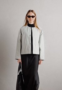 Lys grå, quiltet jakke med rund hals og trykknapper, kombinert med en svart lang kjole og svart håndveske. Strømlinjeformet, moderne design.