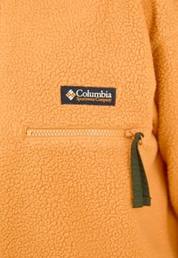 Orange fleecejacka med en ficka med dragkedja och en svart etikett från Columbia Sportswear Company. Strukturerad, varm material med en mjuk finish.