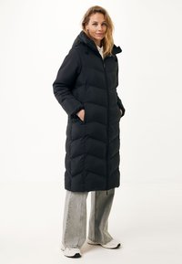 Černá plněná bunda s kapucí, s prošívaným designem, předním zipem a bočními kapsami. V kombinaci s světle šedými kalhotami s širokými nohavicemi.