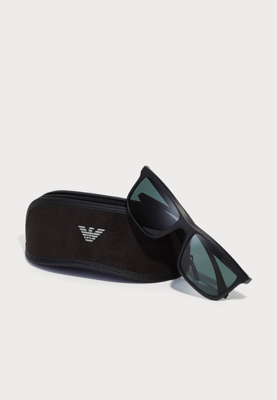Ochelari de soare negri cu lentile întunecate, având un finisaj mat și un design sportiv, împreună cu o husă moale neagră cu un logo.