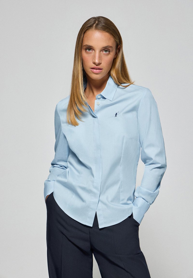 Ljusblå skjorta med knappar av mjukt tyg, med klassisk krage, långa ärmar och en liten logotyp på bröstet.