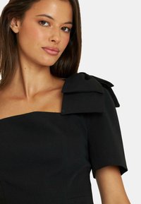 Schwarzes Kleid mit quadratischem Ausschnitt und kurzen Ärmeln. Verfügt über große Schleifenakzente an den Schultern, aus glattem Stoff mit einer strukturierten Form.