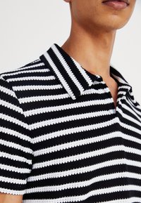 Czarna i biała koszulka polo w paski z teksturowanej tkaniny, krótkimi rękawami i płaskim kołnierzykiem. Zbliżenie skupiające się na szczegółach dekoltu.