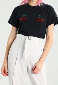 Svart bomullströja med röda körsbärsapplikationer på bröstet; kombinerad med höga vita randiga byxor och ett bälte.