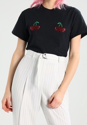 Camiseta de algodón negra con apliques de cereza roja en el pecho; combinada con pantalones de pinzas blancos de tiro alto y un cinturón.