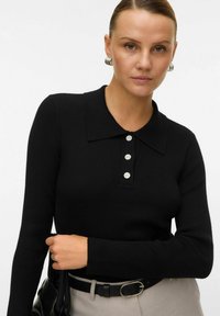 Must kui viigiga polo särk mustades ribades, klassikalise kraega, millel on kaeluses kolm dekoratiivset lillelist nuppu. Rõhutatakse tekstuuri ja lihtsust.