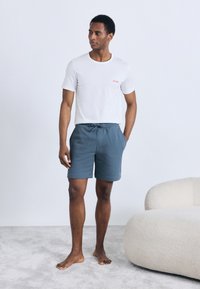 White cotton t-shirt with red logo and blue check-patterned shorts. Model is barefoot, standing on a light carpet next to a rounded cream sofa.