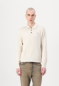Gräddfärgad långärmad pikétröja med knäppning, ribbade ärmslut och en liten logotyp på vänster bröst. Stylerad med ljusa denimjeans.