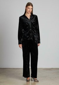 Camisa de botón de terciopelo negro con un diseño de frente retorcido, combinada con pantalones sueltos a juego, que presentan un brillo sutil y una textura suave.