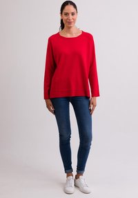 Maglione rosso a maniche lunghe con scollo a barca e orlo a costine, abbinato a jeans skinny blu scuro e sneakers bianche.