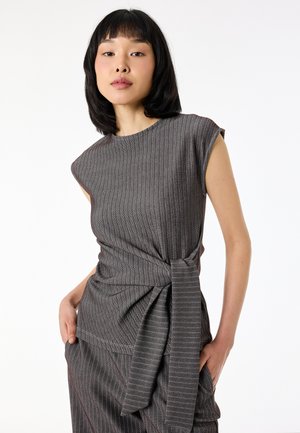 Femme portant un haut gris sans manches à fines rayures avec un nœud sur le côté et un pantalon assorti, posant les mains dans les poches sur fond blanc.