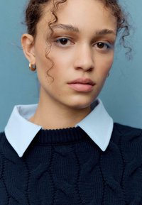 Maglione blu navy a trecce con camicia colletto azzurro chiaro. Modello con capelli ricci, luce soffusa, trucco neutro e orecchini a cerchio dorati.