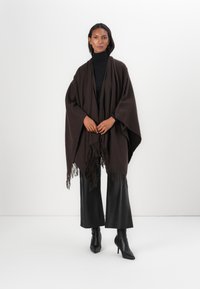 Brauner Poncho mit lockerem Fall, fransigen Kanten, hergestellt aus weichem Stoff. Kombiniert mit einem schwarzen Rollkragenpullover, Lederhosen und schwindelerregenden Stiefeln.