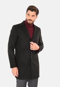 Cappotto di lana nero con chiusura a un bottone, rever a scalpello e due tasche frontali. Indossato sopra un maglione a collo alto borgogna.
