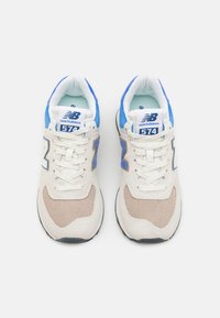 Tenisky v krémové a světle hnědé barvě s modrými akcenty, materiály z mesh a semiše, výrazné logo "New Balance" a design se šněrováním.