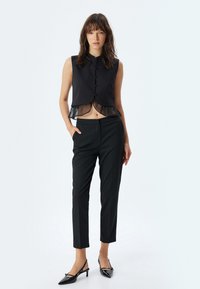 Zwarte cropped vest met een mandarin-kraag, knopen aan de voorkant en een doorschijnende ruchezoom, gecombineerd met op maat gemaakte zwarte broeken en puntige hakken.