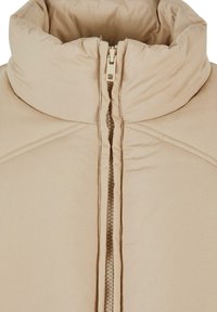 Beige pufferjakke med høy krage, frontglidelås og quiltet tekstur. Har sydd sømmer og mykt, polstret materiale.