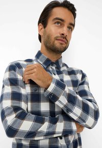 Camicia a maniche lunghe a quadri nei colori bianco, blu marino e marrone; presenta un colletto con bottoni e polsini con due bottoni; texture del tessuto morbida.