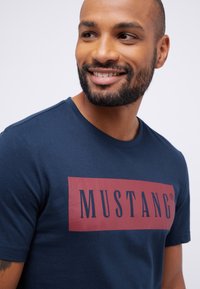 Tamsiai mėlynas medvilninis marškinėlis, kurio priekyje yra raudonojo vyno spalvos stačiakampis su užrašu "MUSTANG." Trumpomis rankovėmis ir apvaliu kaklu, su glotnia tekstūra.