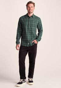 Green plaid button-up shirt with long sleeves, chest pocket, and curved hem, paired with black pants and black sneakers with white accents.
