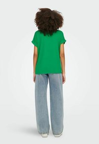 Personne aux cheveux bouclés, vue de dos, portant une chemise verte à manches courtes, un jean bleu à jambes larges et des chaussures blanches sur un fond uni.