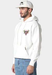 Uomo che indossa una felpa bianca con un piccolo grafico colorato sul petto e un cappellino bianco con testo nero, abbinato a jeans blu, rivolto a sinistra.