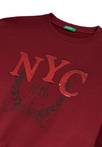 Felpa borgogna con lettere "NYC" in paillettes rosse e testo nero "Girl Team Sisterhood" circondata da un design a ghirlanda d'alloro.