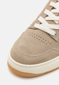 Beige suede kenkä, jossa on rei'itetty kärki, litteät kermaiset nauhat ja valkoinen kumipohja, jossa on vaaleanruskea strukturoitu ala.