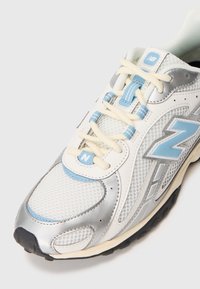 Sneaker bianco con accenti argento, dettagli azzurri e tomaia in rete. Presenta lacci rotondi e una suola in gomma texture. Progettato per il comfort.