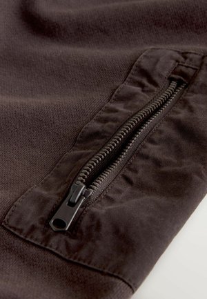 Tmavě hnědá látka s hladkým texturem, s výrazným zipem na straně. Zip je černý s viditelnými zuby.