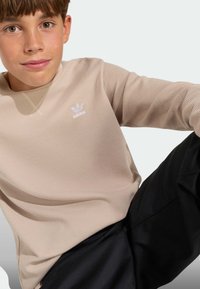 Lahka bež dolgo rokavno majico z V-izrezom in belim adidas logotipom na prsih, kombinirano s črnimi hlačami.