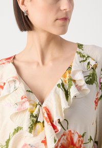 Blusa floreale color crema con scollatura arricciata; presenta motivi floreali colorati, tessuto leggero e un dettaglio colletto a forma di V.