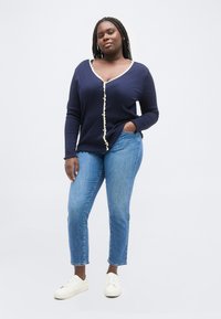 Γυναίκα με κοτσίδες, που φοράει μπλε σκούροπλεκτή ζακέτα με κουμπιά και άσπρη μπορντούρα, ανοιχτό μπλε τζιν και άσπρα αθλητικά παπούτσια, στέκεται με το ένα χέρι στην τσέπη.