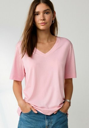 Ung kvinde med langt brunt hår iført sølv hoop øreringe, lyserød V-hals T-shirt, blå jeans og sølvur, hænderne i lommerne.