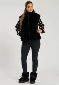 Falsk päls väst i svart med hög krage, parat med randiga långärmade tröjor. Modellen bär svarta leggings och kraftiga svarta stövlar med pälsdetaljer.